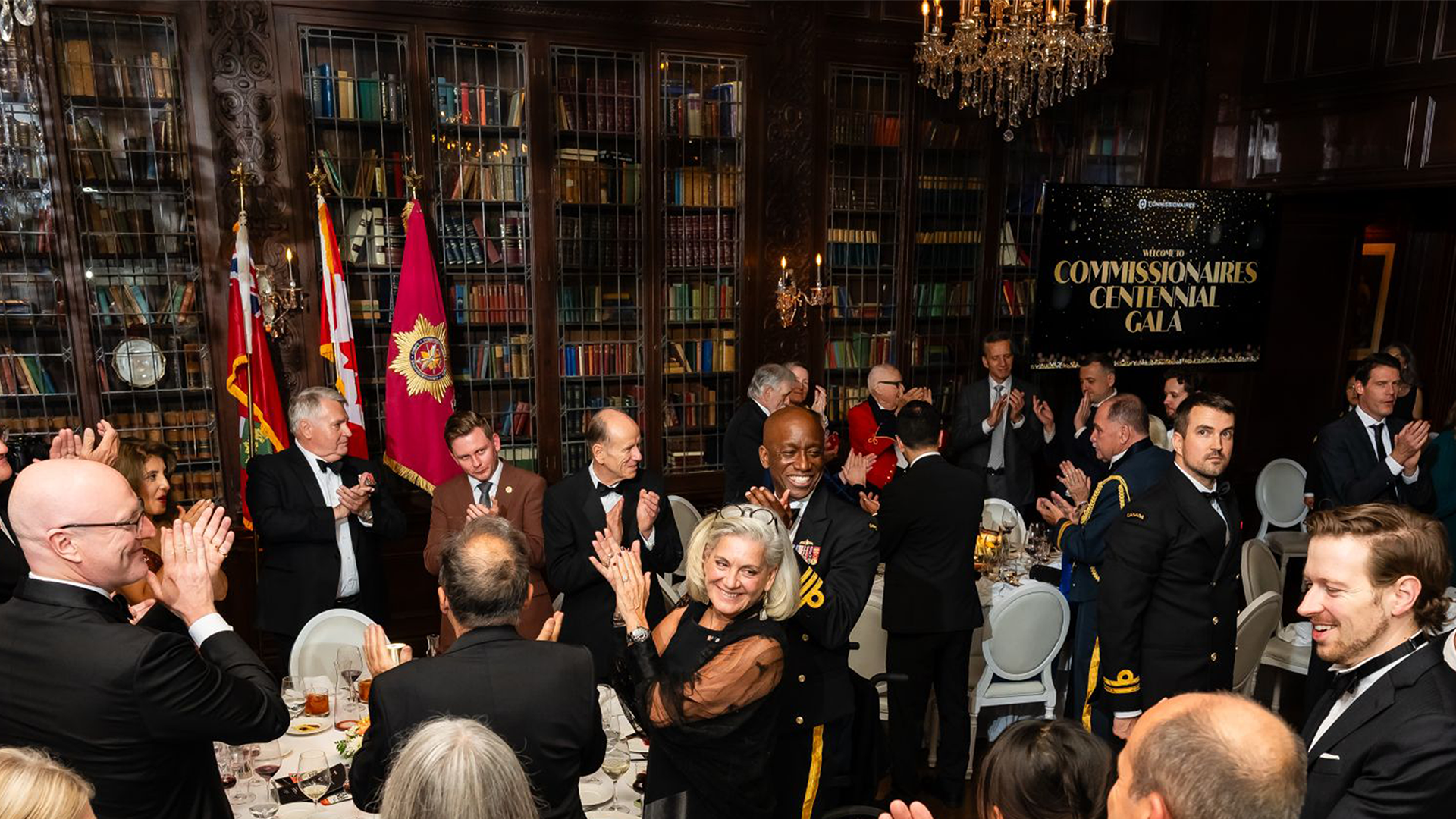 Gala photo showing attendees socializing, with a TV screen in the background displaying the black-and-gold Centennial Gala design.