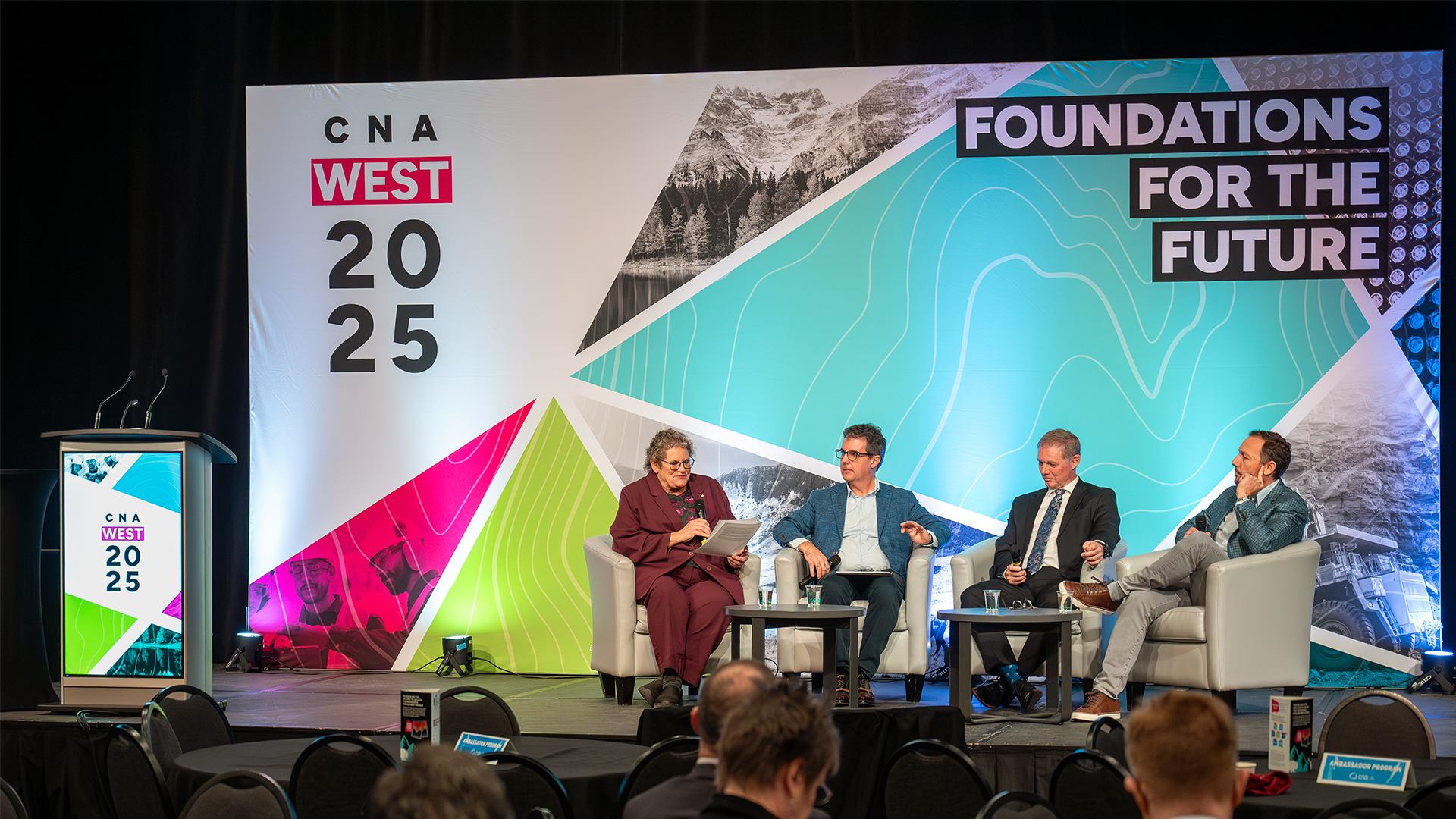 Photo from CNA West 2025 showing the stage backdrop in use: four people seated on stage right, and a podium on stage left.